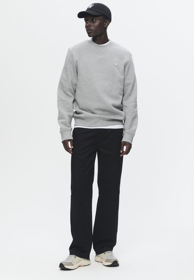 Hall halliga sweatshirt ümmarguse kaelusega ja väikse logoga, koos mustade pükstega. Mees kannab musta mütsi ja beeže sneakersite, millel on hallid aktsendid.