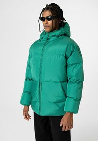 Groene gewatteerde jas met een capuchon, met een rits aan de voorkant, elastische manchetten en een gladde, glanzende afwerking. Draagt over donkere broeken.