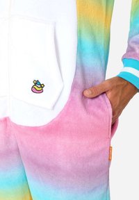 Rainbow-gradient onesie with soft, plush fabric; features a white front pocket and an embroidered colorful logo. Cuffs are light blue with stripes.