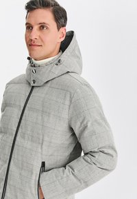 Jasnoszara pikowana kurtka w kratę, z wysokim kołnierzem, regulowanym kapturem i czarnym zamkiem błyskawicznym. Materiał wygląda na gładki i ciepły.