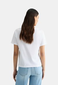 T-shirt bianca a maniche corte con una vestibilità rilassata, realizzata in tessuto morbido. La vista posteriore mostra un collo rotondo e cuciture, abbinata a jeans azzurri chiari.