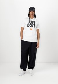 Balta marškinėliai su juodu užrašu "JUST DO IT" storais rašmenimis ir oranžiniu Nike logotipu, derinami su laisvomis juodomis kelnėmis ir baltomis sportinėmis bateliais. Juoda kepurė.