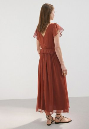 Femme debout, portant une robe midi fluide couleur rouille avec des manches papillon et des sandales plates noires à brides, vue de profil sur un fond uni.