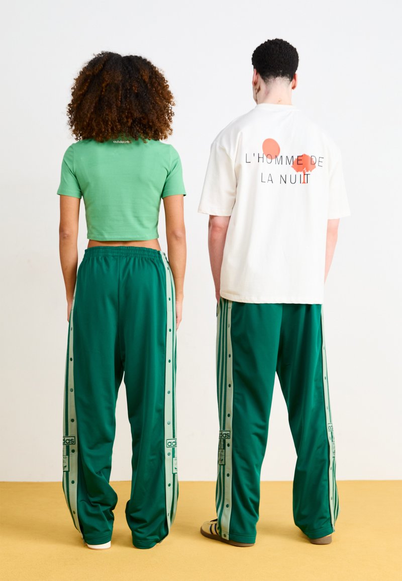 Top corto verde, camisa blanca de gran tamaño con gráfico, pantalones de chándal verdes con botones laterales y rayas, todo usado por dos personas de espaldas.
