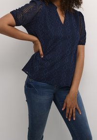 Mörkblå blus med texturerad tyg med diamantmönster, korta puffärmar och V-ringning. Kombinerad med figurnära blå jeans.