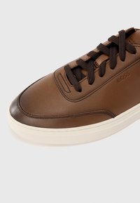 Brune lærersneaker med rund tå, mørkebrune snørebånd og kremfarget gummisåle. Har subtil merkevare og teksturert overflate.