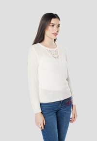 Witte gebreide trui met lange mouwen, kantdetails bij de halslijn en een aangesloten pasvorm. Draagt een blauwe spijkerbroek met kleurrijke borduursels.