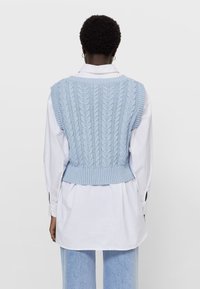 Osoba z krótkimi, kręconymi włosami ma na sobie jasnoniebieski bezrękawnik w warkocze, założony na białą koszulę z długim rękawem oraz jasnoniebieskie spodnie, odwrócona plecami.