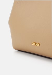 Beige skinnveske i pebble-lær med teksturert overflate, med en gull DKNY-logo på forsiden og en strukturert, vinklet form.