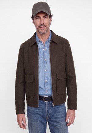 Un homme porte une veste zippée marron foncé par-dessus une chemise en denim bleu et un jean. Il porte une casquette foncée sur un fond blanc uni.