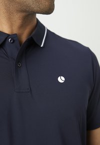 Vyras vilki tamsiai mėlyną polo marškinėlį su dviem mygtukais, balto apykakle ir mažo balto apvalaus logotipo kairėje krūtinės pusėje.