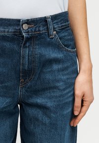 Μπλε τζιν παντελόνι από denim με κλασικό σχέδιο πέντε τσεπών, ραφές σε χαλκό, κλείσιμο με κουμπιά μπροστά και μαλακή, υφασμένη υφή.