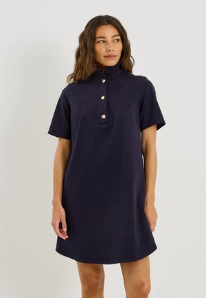 Mujer con cabello ondulado que lleva un vestido azul marino de manga corta con cuello con volantes y cuatro botones en forma de corazón, de pie frente a un fondo liso.