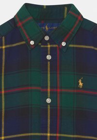 Kostkovaná košile na knoflíky v zelené, námořnické a červené barvě, z flanelu. Má klasický límec, bílé knoflíky a zlaté logo na hrudi.