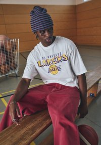 Bijela majica s grafikom "Los Angeles Lakers" u ljubičastoj i žutoj boji, uparena s širokim crvenim trapericama. Nosena plavo-ljubičasta prugasta kapa.