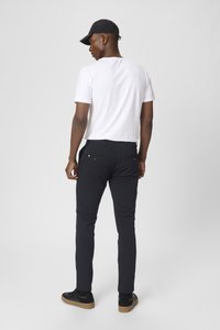 Pantalones ajustados negros con bolsillos laterales y traseros, combinados con una camiseta blanca. El modelo lleva zapatillas negras y una gorra negra.