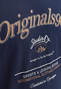 Tums zilā džersija ar lieliem bežiem burtiem "Originals90" un mazākiem tekstiem. Iekļautas kalnu grafikas un izliekta balta līnija.