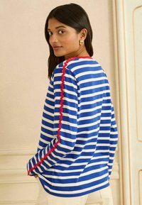 Blau-weiß gestreiftes Langarmoberteil mit roten, muschelförmigen Akzenten an der Schulternaht, mit lockerem Schnitt und Rundhalsausschnitt.