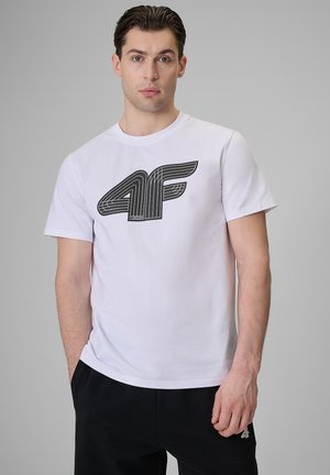 Młody mężczyzna z krótkimi ciemnymi włosami, ubrany w białą koszulkę z czarnym logo "4F" i czarne spodnie, stoi na jasnoszarym tle.
