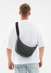 Borsa a tracolla nera realizzata in materiale morbido con un design curvo, cinturino regolabile e logo in rilievo discreto sul davanti.