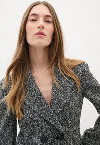Houndstooth raksts, dubultā krusta žakete melnbalti ar lielām melnām pogām, ar dziļu V veida kaklu un teksturētu audumu.