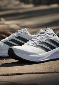 Paire de chaussures de course blanches avec des rayures noires reposant sur une surface pavée sous un éclairage naturel extérieur.