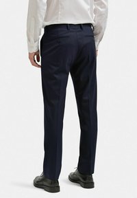 Marina blå kostymbyxor tillverkade av slätt tyg med skräddarsydd passform. Två bakfickor med knappstängning; rak byxstil. Bärs med svarta skor.