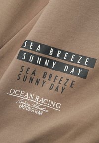 Un tessuto marrone con una texture liscia presenta un testo nero con la scritta "SEA BREEZE SUNNY DAY" e "OCEAN RACING Sailing Adventure" in bianco.