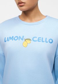 Άτομο που φοράει ανοιχτό μπλε φούτερ με το κείμενο "LIMON CELLO" και δύο κίτρινα λεμόνια με πράσινο φύλλο τυπωμένα ανάμεσα στις λέξεις.