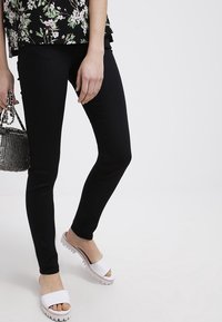 Černé elastické legíny s přiléhavým střihem, doplněné o bílé nazouvací sandály s texturovanou podrážkou. Doplňky zahrnují malou stříbrnou kabelku.