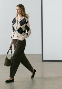 Vrouw die binnenshuis loopt, gekleed in een zwart-beige argyle vest, beige top, donkere losse broek, zwarte ballerina's, en een donkerbruine handtas draagt.