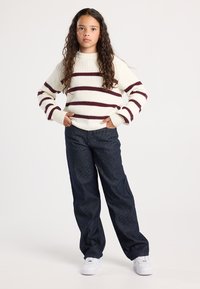 Tyttö, jolla on pitkät kiharat hiukset, seisoo kädet taskuissa, päällään kermansävyinen neule burgundinvärisin raidoittein, tummat raidalliset housut ja valkoiset lenkkarit.