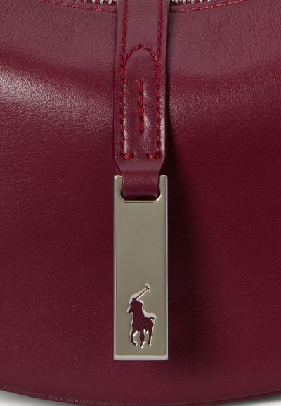 Geantă din piele bordeaux cu textură netedă, având un etichetă metalică cu un emblema de jucător de polo. Căptușelile sunt în nuanță potrivită. Închizătoare cu fermoar.