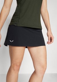 Falda deportiva negra con textura perforada, aberturas laterales y un logotipo discreto a la izquierda. Combinada con una parte superior de color verde oscuro.