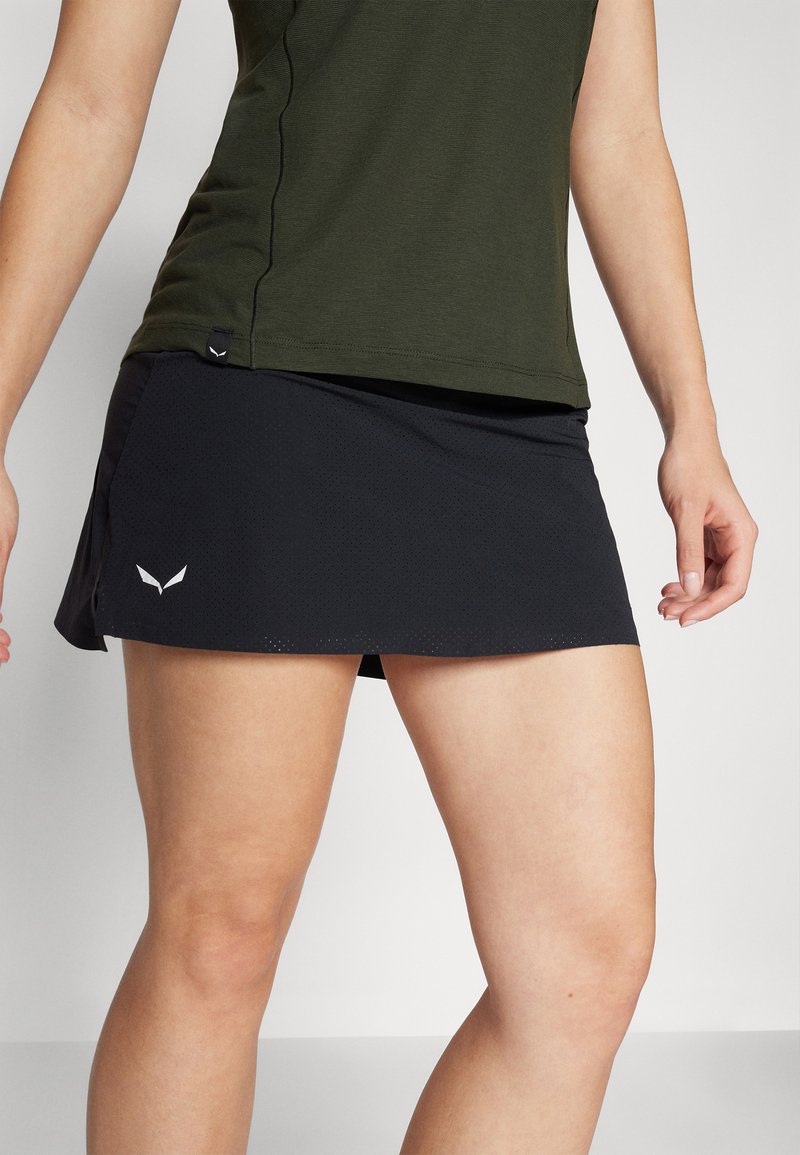 Czarna spódnica sportowa z perforowaną fakturą, bocznymi rozcięciami i dyskretnym logo po lewej stronie. Zestawiona z ciemnozieloną bluzką.