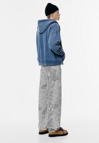 Felpa blu in pile con tasca a marsupio, abbinata a pantaloni grigi pixelati e scarpe marroni con suole nere e lacci blu.