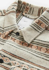 Tkaná flanelová košile v béžové barvě s černými a měděnými vodorovnými pruhy a vzorovanými akcenty. Má klasický límec a černé knoflíkové detaily.