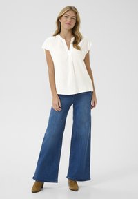 Bluză albă cu mânecă scurtă, decolteu în V și detalii cu pliuri, asortată cu blugi blue denim cu picioare largi și bocanci maro până la gleznă.