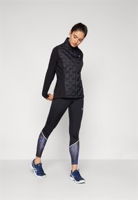 Black padded jacket with a high collar, paired with black leggings featuring a colorful print and white accents. Blue athletic shoes complete the outfit.