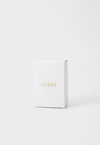 Biały prostokątny box z złotym logo "GUESS" umieszczonym na środku przodu, na tle gładkiego białego tła.