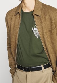 Braune Zip-Jacke mit strukturiertem Finish über einem grünen T-Shirt mit einem weißen Grafikdesign. Schwarzer Gürtel mit silberner Schnalle und beige Hose.