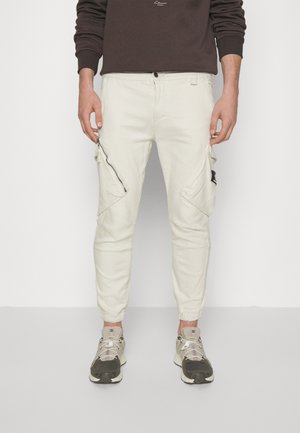 Mężczyzna w kremowych spodniach typu cargo z zamykanymi na zamek kieszeniami po bokach i szarymi trampkami, stojący na tle jednolitego białego tła.