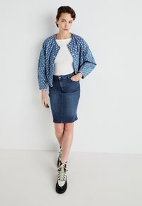 Gequilted blauwe denimjacket met wit bloemenpatroon, gedragen over een witte top en een donkere denim kokerrok, gecombineerd met stevige zwart-witte laarzen.