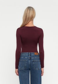 Una donna con lunghi capelli biondi indossa un top aderente a maniche lunghe color bordeaux infilato in jeans blu a vita alta, in piedi contro uno sfondo bianco.