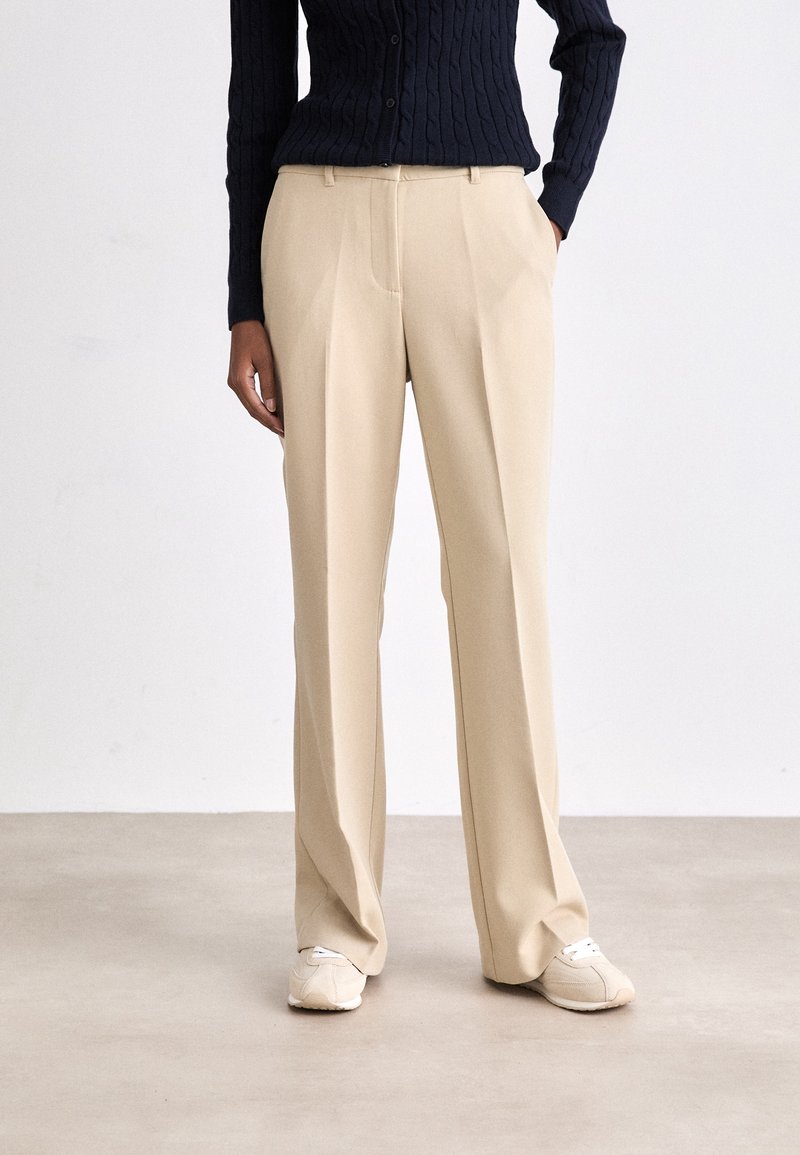 Personne portant un pantalon beige à jambes larges avec poches latérales, des baskets blanches et un pull en maille noir à manches longues sur un fond uni.