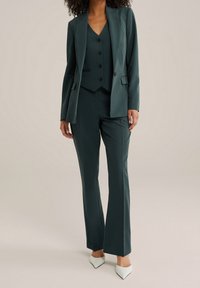 Grön skräddarsydd kostym med en figursydd kavaj med slag, en väst med tre knappar och utsvängda byxor, parat med vita spetsiga klackar.