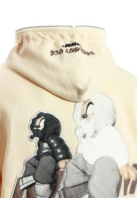 Sudadera beige con cremallera en la capucha, que presenta personajes de dibujos animados bordados que llevan chaquetas con capucha en blanco y negro.