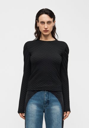 Kvinde med mørkt hår iført tekstureret sort langærmet top og højtaljede blå jeans med sorte buede accenter, står mod en hvid baggrund.