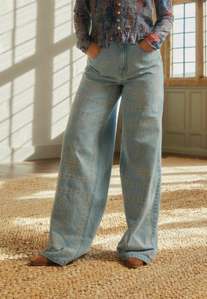 Person trägt eine weit geschnittene Jeans mit bestickten geometrischen Mustern und ein mehrfarbiges, mit Knöpfen versehenes Oberteil und steht auf einem gewebten Teppich neben einem Fenster.