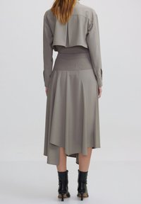 Frau in einer taubfarbenen, verkürzten Jacke und asymmetrischem Midirock, kombiniert mit schwarzen, hohen Stiefeletten, von hinten gesehen.